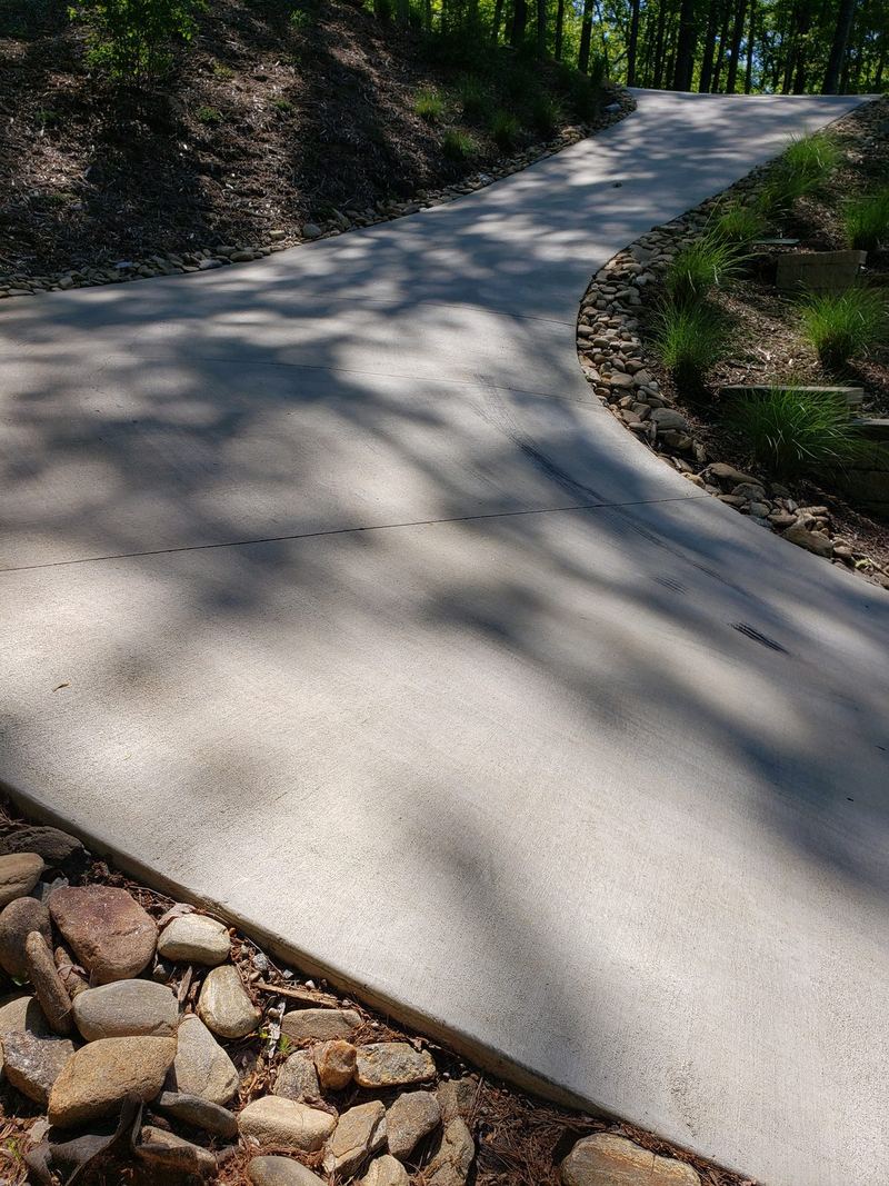 Curved concrete walkway edged with river rock winding through a wooded yard